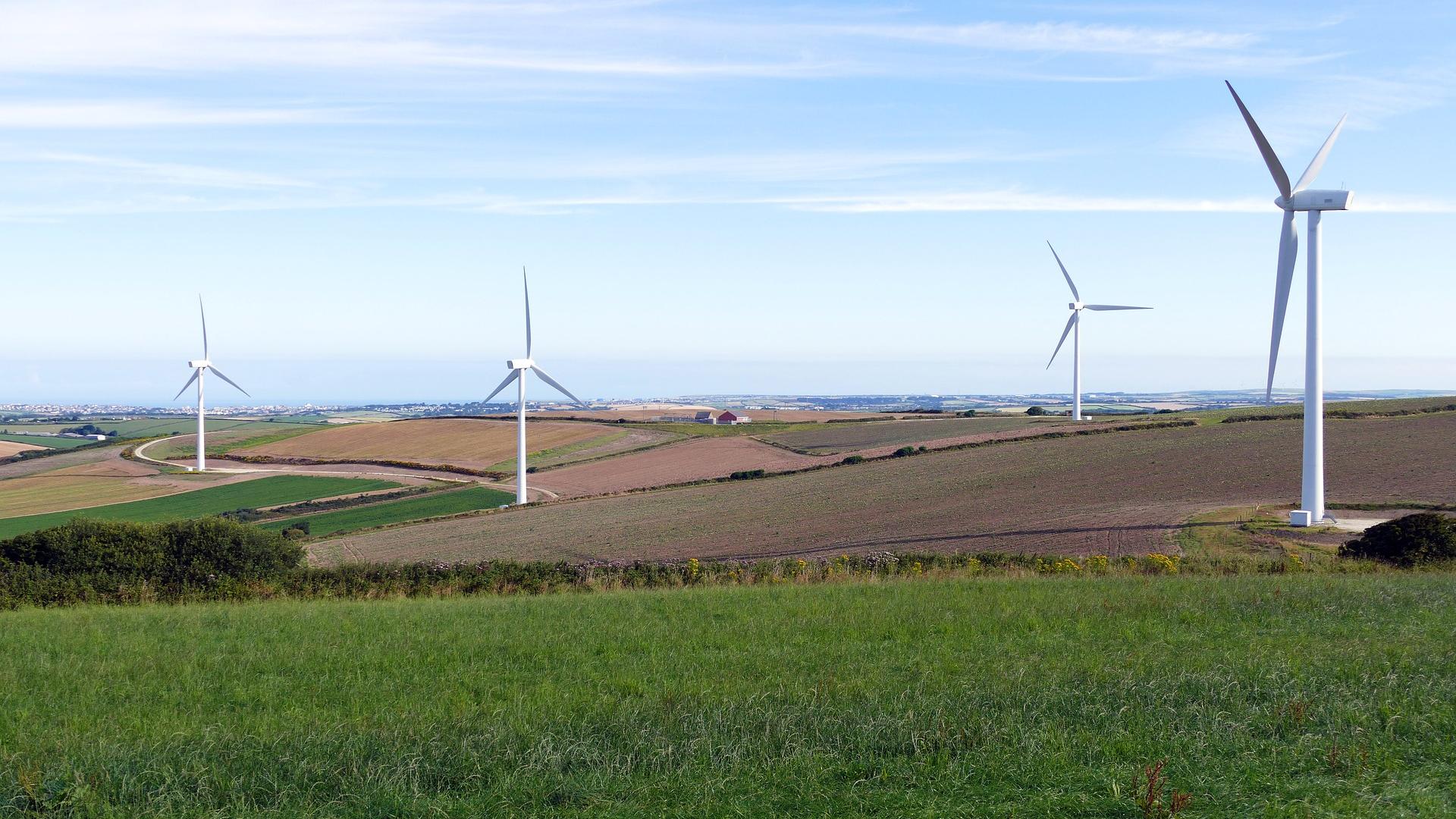 Een afbeelding van vier windturbines tussen landbouw akkers, achter de velden ligt een stad